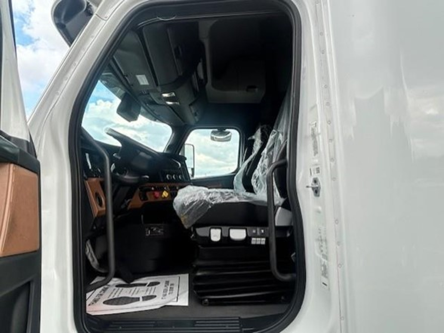 Truck cab interior view of a 2023 Freightliner Cascadia 126 from the driver’s side door, showing the steering wheel, dashboard, and driver seat wrapped in plastic.