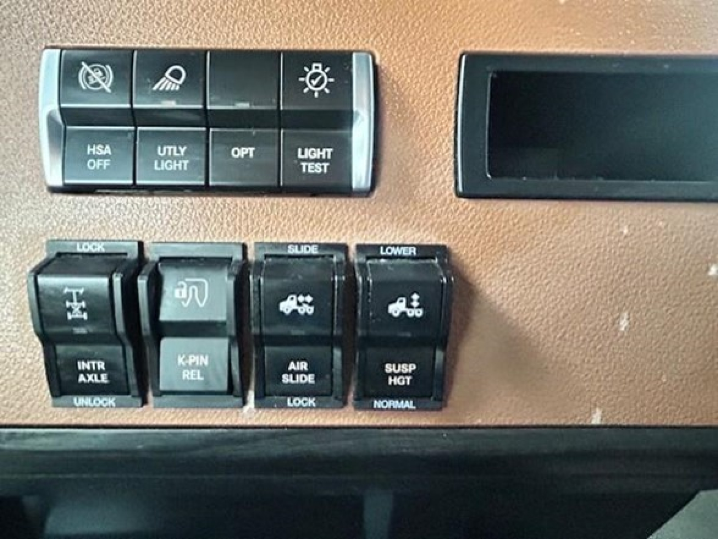 Dashboard control panel of Freightliner Cascadia showing switches labeled HSA OFF, UTLY LIGHT, OPT, LIGHT TEST, INTR AXLE, UNLOCK, K-PIN REL, AIR SLIDE, SUSP HGT, LOWER, and other icons.