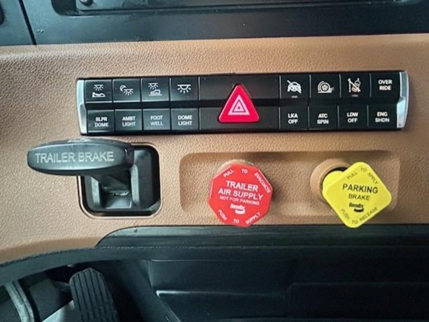 Dashboard panel of a Freightliner truck showing trailer brake control, red hazard switch, and yellow parking brake knob with various indicator icons above.