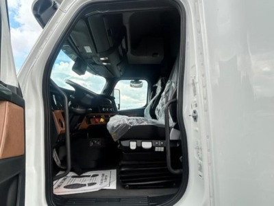 Truck cab interior view of a 2023 Freightliner Cascadia 126 from the driver’s side door, showing the steering wheel, dashboard, and driver seat wrapped in plastic.