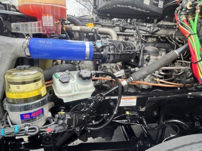Engine bay of a 2023 Freightliner Cascadia 126 showing coolant reservoir, blue silicone hose, yellow canister, various wiring, and components