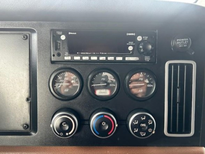 Dashboard panel showing gauges and radio in a Freightliner Cascadia cab. Visible: radio head unit, three circular gauges, climate controls, and a power outlet on the right.