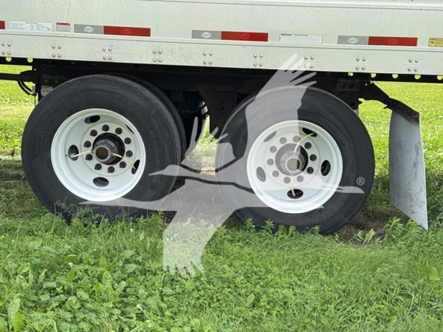 Two white rear trailer wheels with dual tires under a flatbed trailer; visible rims, tires, and a portion of the undercarriage with grass beneath.