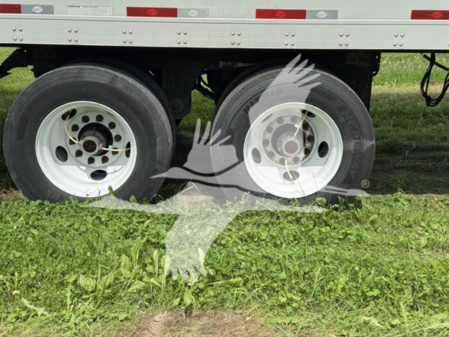 Dual trailer wheels with white rims under a Utility trailer, with grass and a watermark of two hands image overlay.