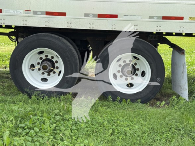 Two white rear trailer wheels with dual tires under a flatbed trailer; visible rims, tires, and a portion of the undercarriage with grass beneath.