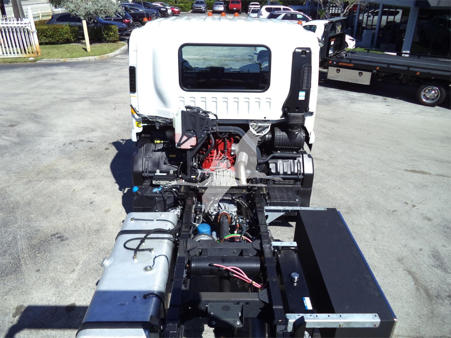 Isuzu rollback truck cab with engine exposed; visible components include red engine, black hoses, silver fuel tanks, and wiring harness; rear flatbed area and surrounding lot.