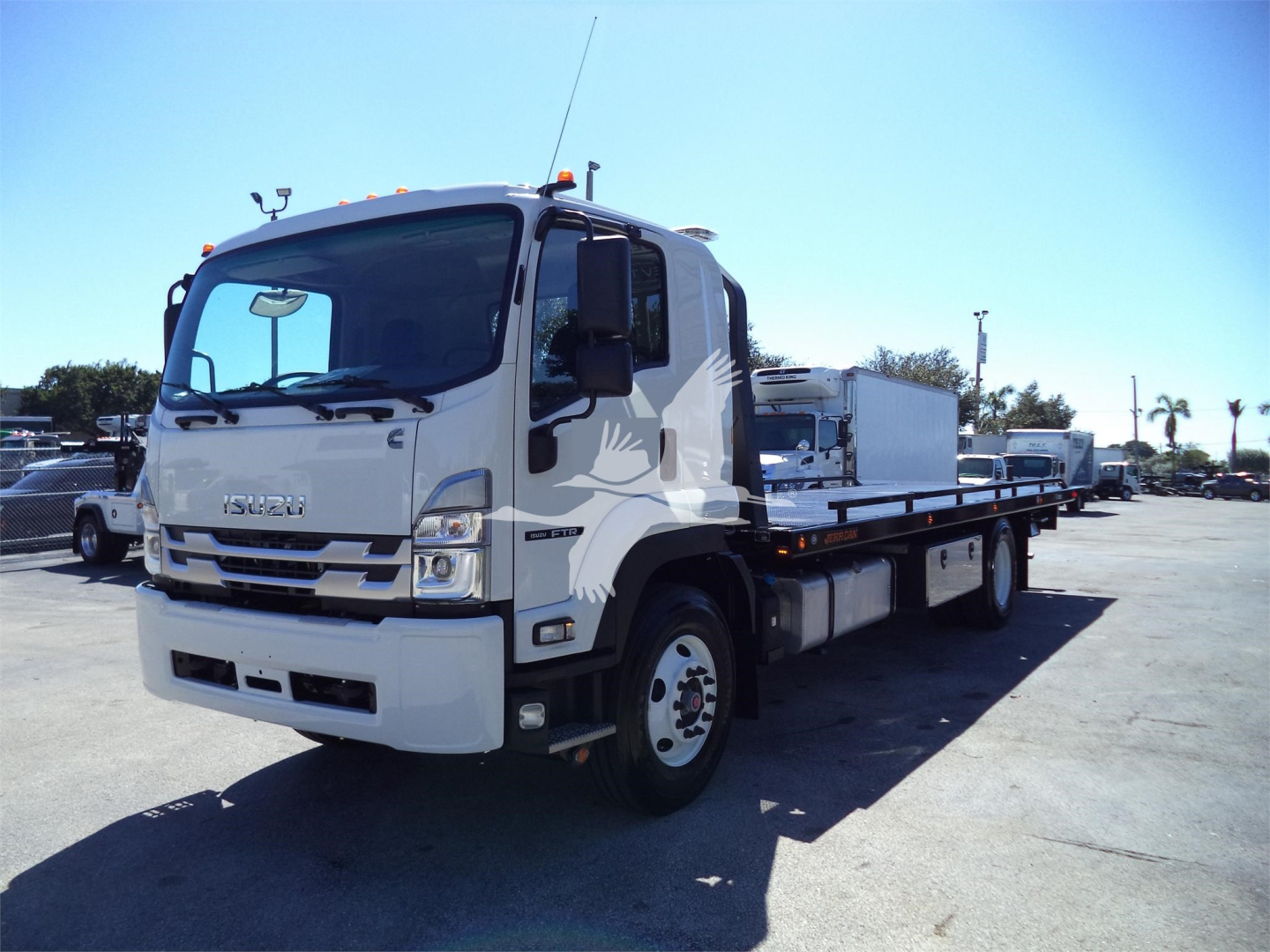 Isuzu flatbed rollback tow truck in a parking lot, cab with Isuzu logo, chassis with long wheelbase, white paint, side mirrors, wrecker equipment not visible