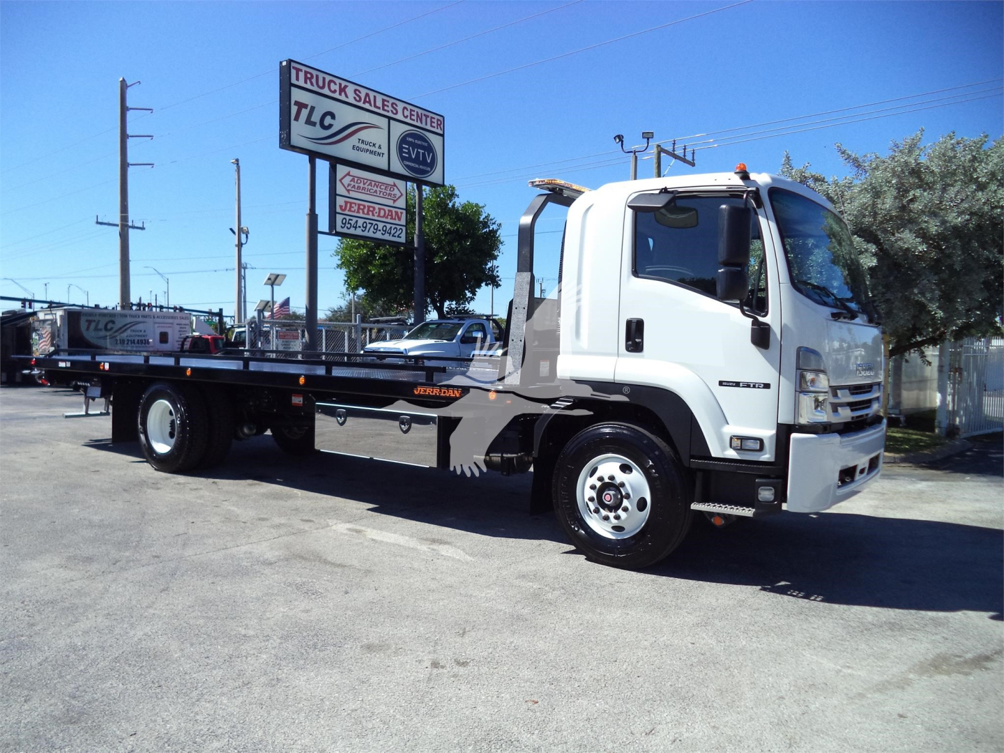 Isuzu FTR rollback truck in side profile view with flatbed, white cab, equipment on bed, dealership lot in background.