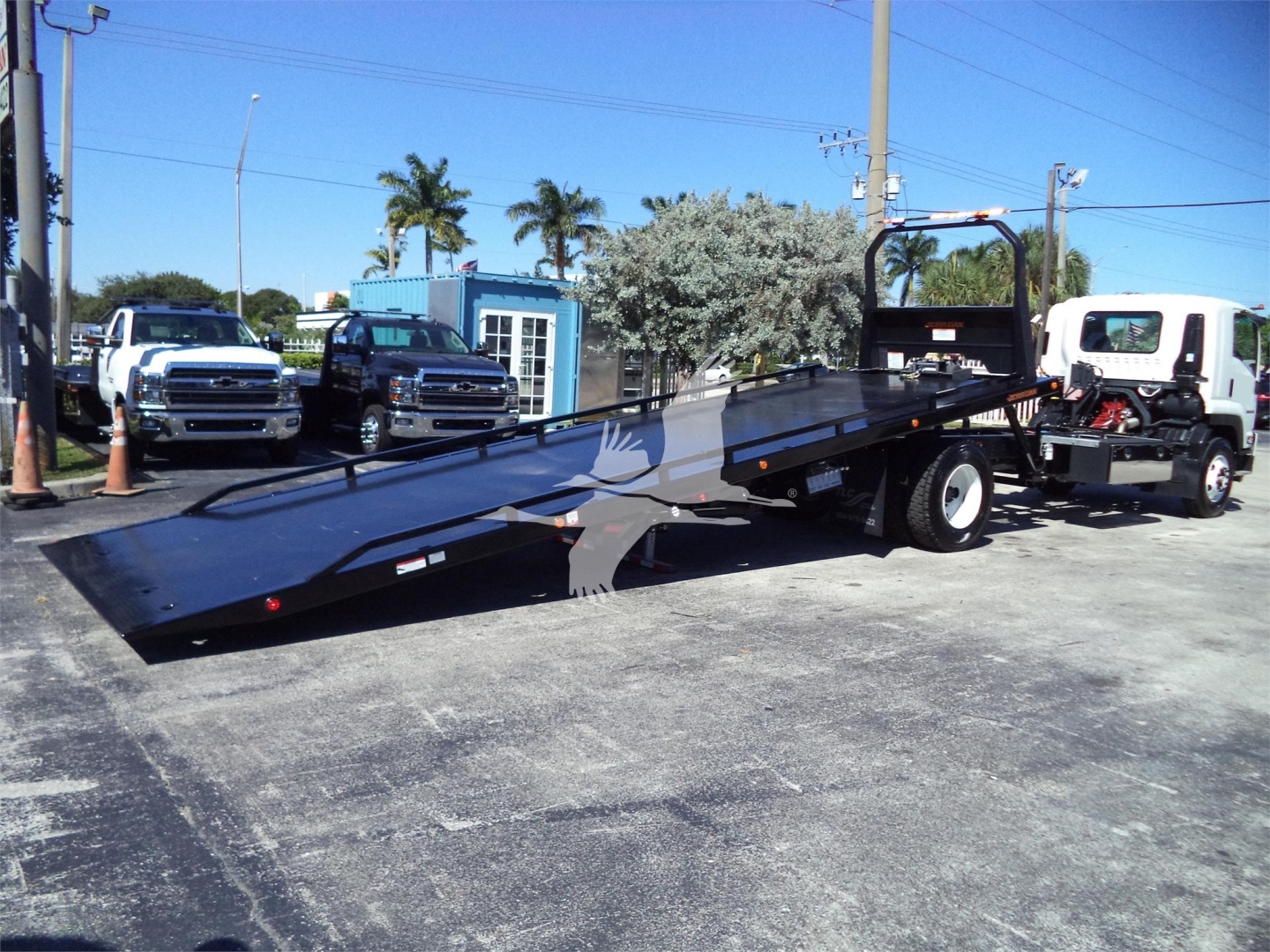Isuzu rollback truck with a long hydraulic trailer ramp deployed, crew cab truck in background, stock/fleet lot.