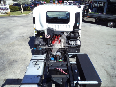 Isuzu rollback truck cab with engine exposed; visible components include red engine, black hoses, silver fuel tanks, and wiring harness; rear flatbed area and surrounding lot.
