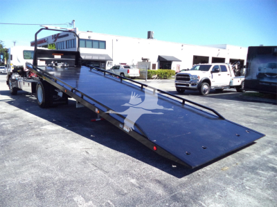 Isuzu FTR rollback tow truck flatbed in service lot showing lowered metal deck with hydraulic mechanism and cab in background.