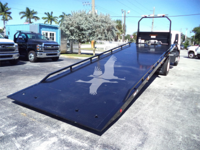 Isuzu FTR rollback tilt bed truck showing a long black low-angle flatbed with a white bird graphic near the front, blue surroundings, and trucks in background.
