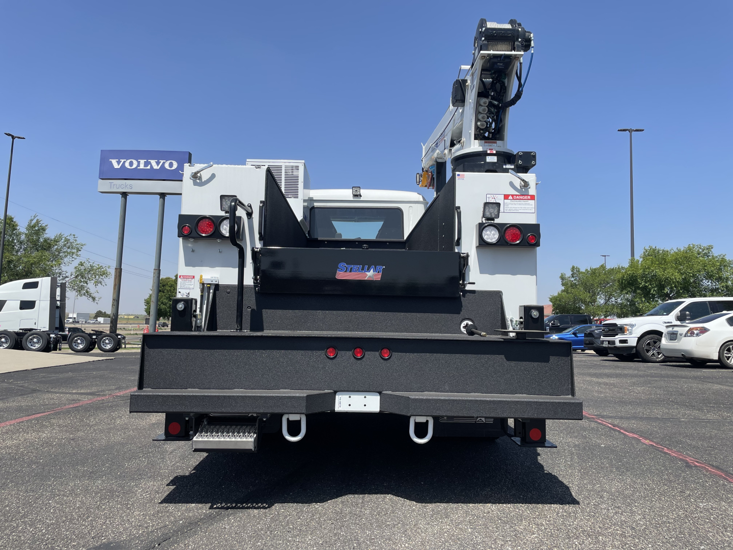 Service/utility truck back view showing a large flatbed, black metal toolboxes, and a mounted crane with control arm on a Mack MD6 chassis. Visible signage and tail lights.