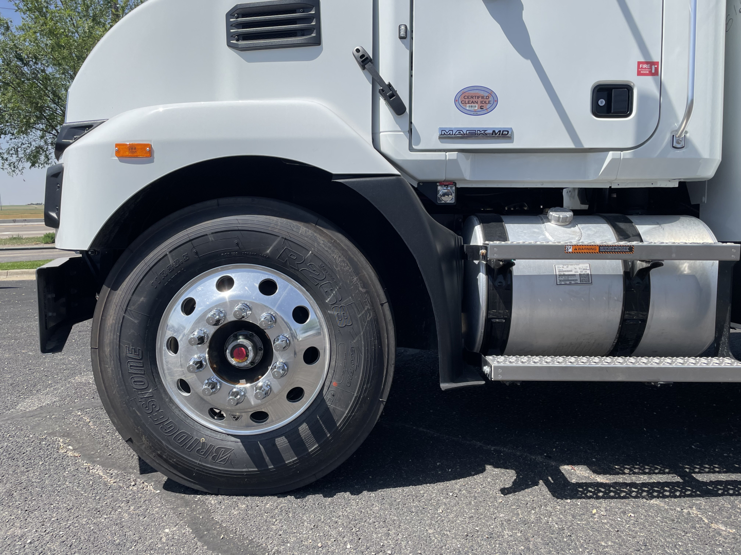 Front-left wheel and lower cab of a 2026 Mack MD6 truck showing tire, rim, fender, steps, and cab doors with badges and a DEF sticker.