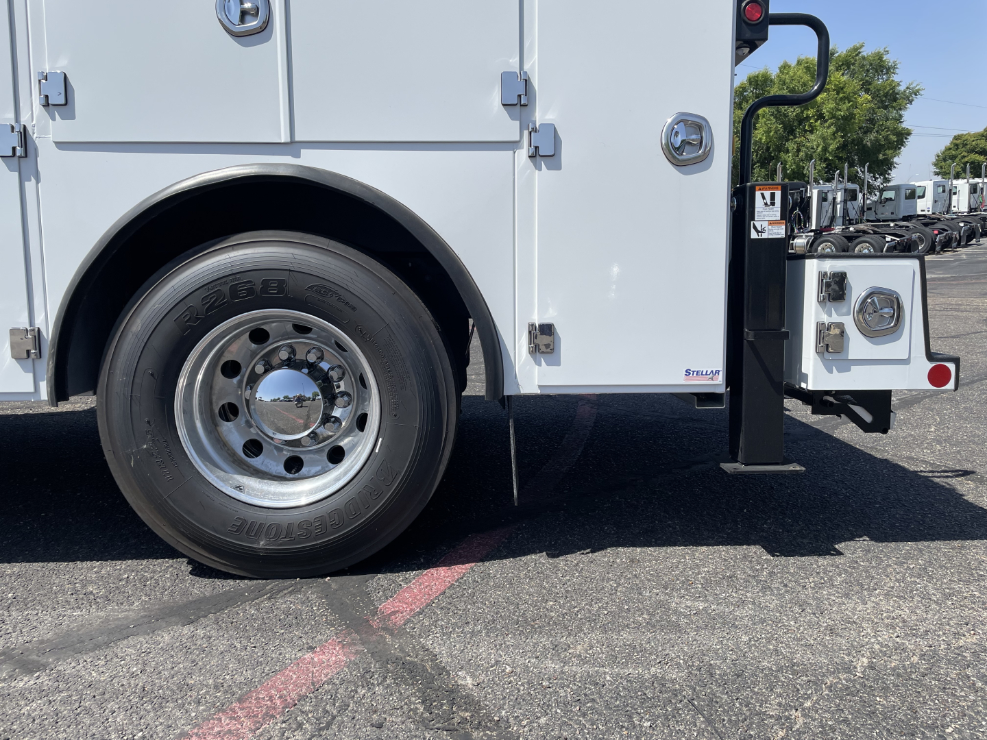 Wheel well of a white Mack MD6 service truck showing tire and chrome hubcap with body panel, lower bumper area, and utility compartments.