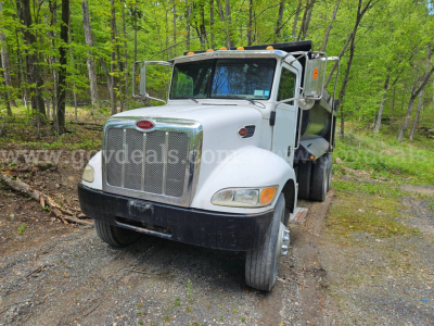 Peterbilt 335 dump truck front three-quarter view in a forested area, white cab with chrome grille, dual rear wheels, dump bed visible.