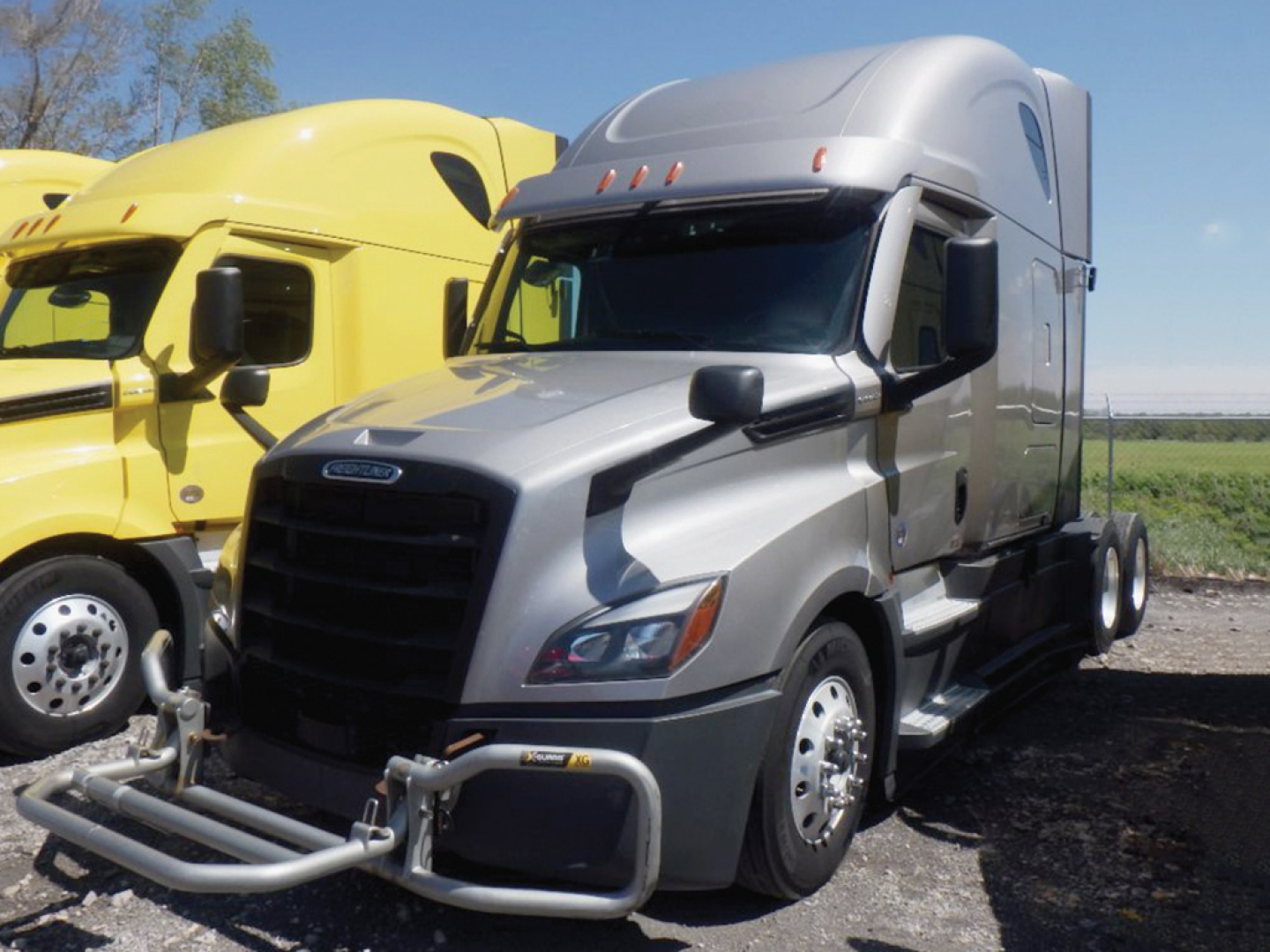 Truck cab of a gray 2022 Freightliner Cascadia126 semi-truck with a silver sleeper, black grille, and front bumper guard, parked next to a yellow truck.