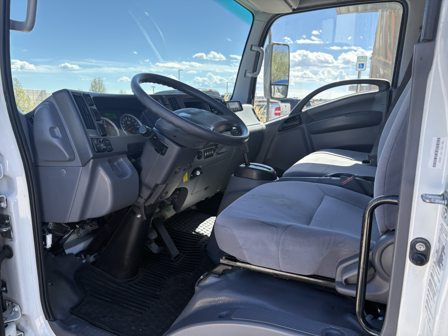 Truck cab interior of a 2020 Chevrolet W4500 with two front seats, steering wheel, dashboard, and manual gear shifter visible; light gray upholstery; large windshield and side mirrors.