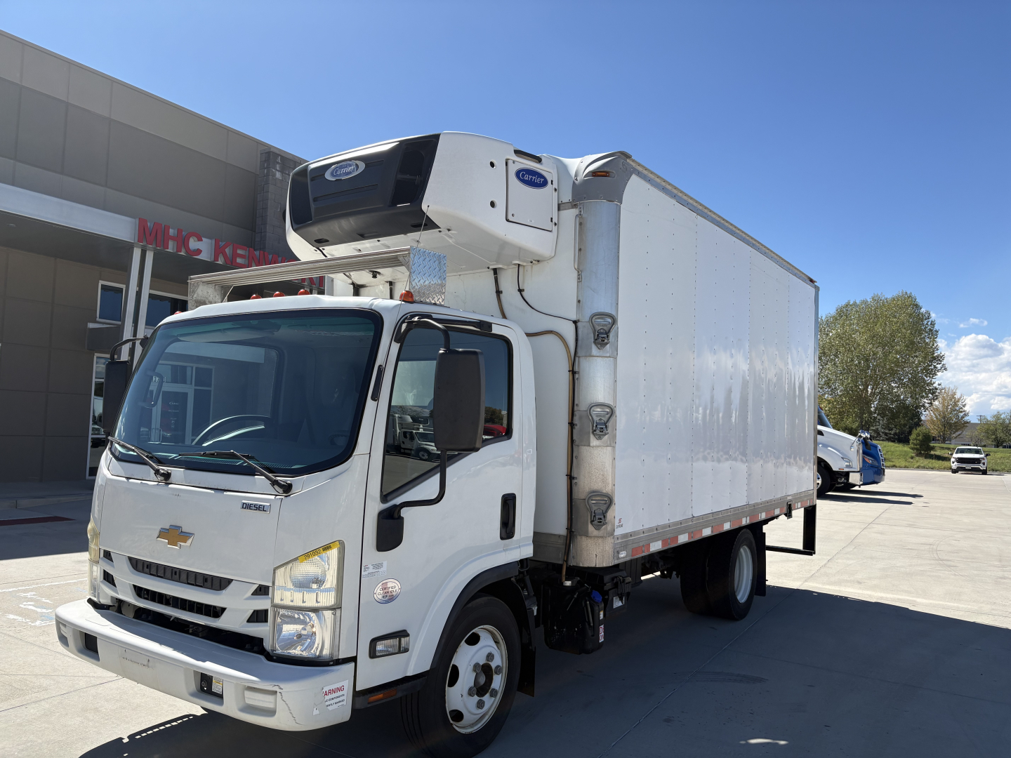 Truck with refrigerated box and front Chevrolet emblem, mounted cold storage unit on cab, white cab and box, refrigeration unit on top front of box, dealership lot in background.