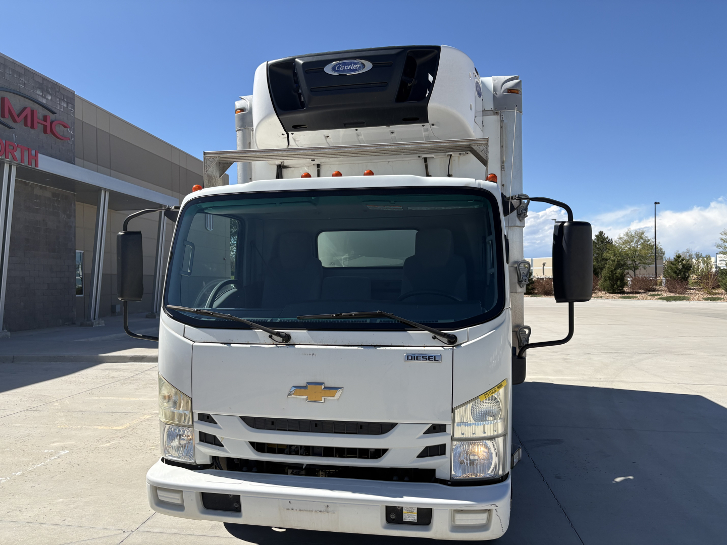 Front view of a white 2020 Chevrolet W4500 refrigerator truck with black side mirrors and a refrigeration unit on the cab roof. Chevrolet emblem on grille, Diesel badge visible near the windshield, large windshield, headlights, and bumper.