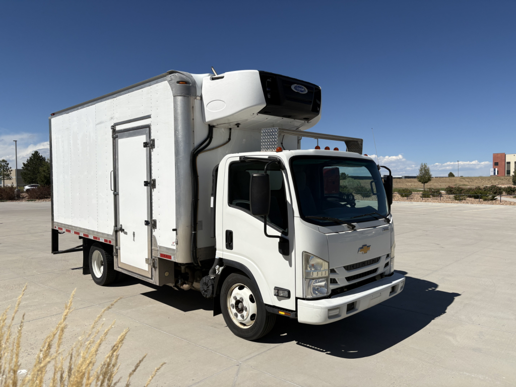 Medium-duty white refrigerated box truck with cargo area and rooftop refrigeration unit, Chevrolet badge on grille, standing on paved lot.