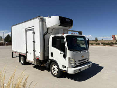 Medium-duty white refrigerated box truck with cargo area and rooftop refrigeration unit, Chevrolet badge on grille, standing on paved lot.