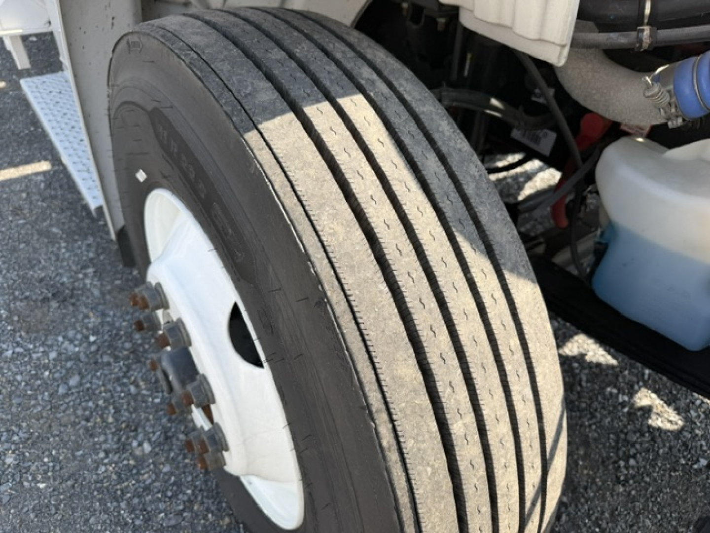 Freightliner M2-106 front wheel area showing dual rear dual tires, white wheel with lug nuts, part of chassis and hydraulic lines.