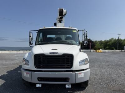 Front view of a white 2023 Freightliner M2-106 crane truck with a mounted crane on top, large black grille, rectangular headlights, and bumper; visible tires and cabin windows.