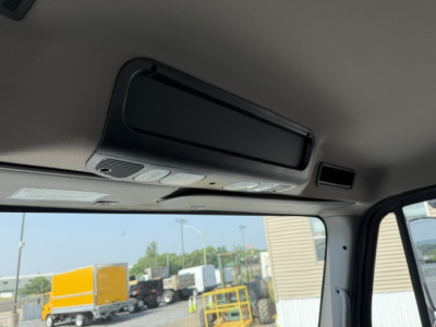 Cab interior overhead console of a Freightliner M2-106 showing sun visor/roof console with controls, light panel, and a window view of yard with trucks.