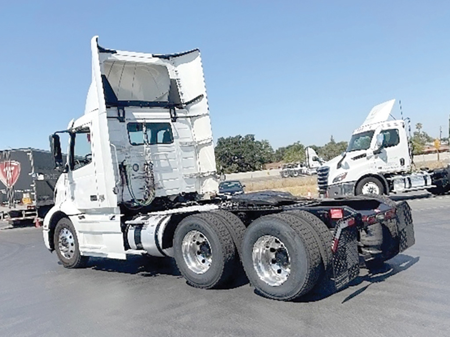 Truck chassis with cab raised, white Volvo VNR 300 daycab over dual rear axles, rear view showing dual tires and fifth wheel, no visible damage, blue sky.