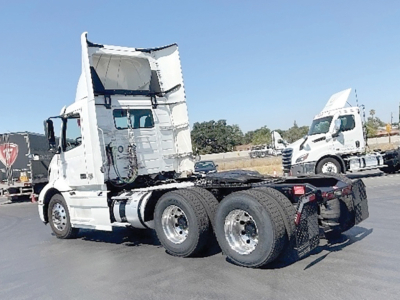 Truck chassis with cab raised, white Volvo VNR 300 daycab over dual rear axles, rear view showing dual tires and fifth wheel, no visible damage, blue sky.
