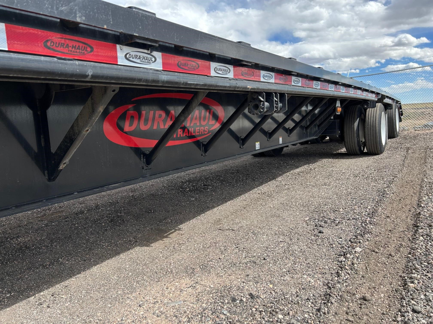 Trailer side showing Dura-Haul Trailers logo and reflective stickers along the top edge; low-profile steel drop deck with dual rear axles and black frame.