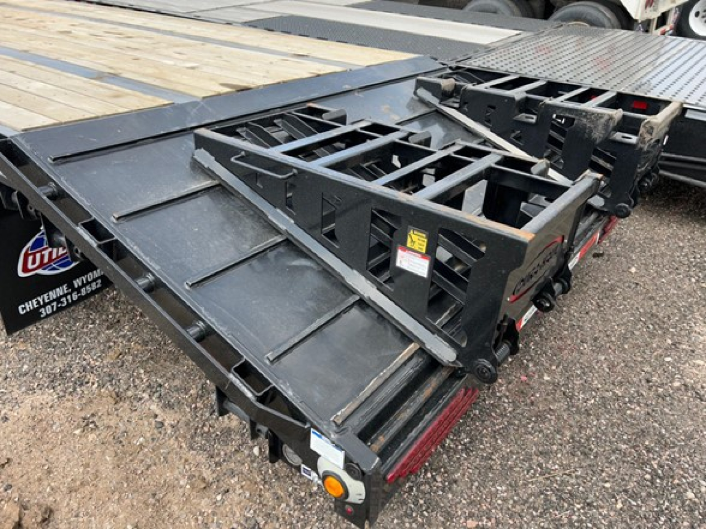 Drop deck trailer in folded position showing steel deck and folding ramp sections with hydraulic or mechanical support; components include hinges, frame rails, and safety stickers.
