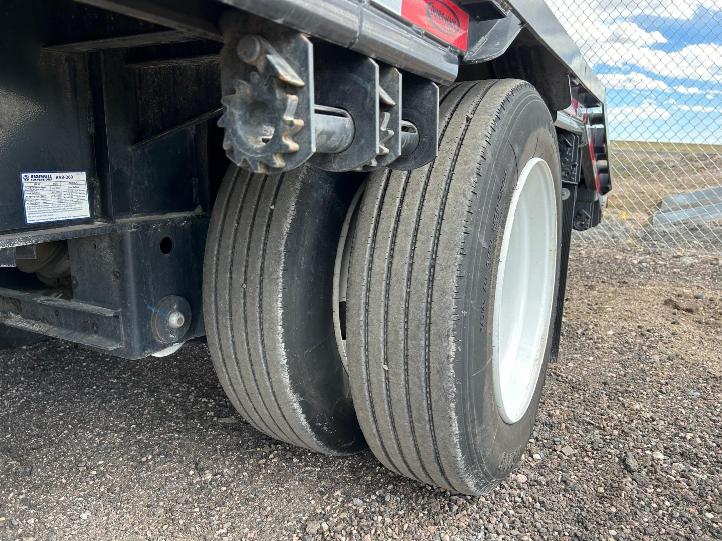 Dual rear tires of a drop deck trailer with black frame and white wheel rims, gravel ground, clear sky.