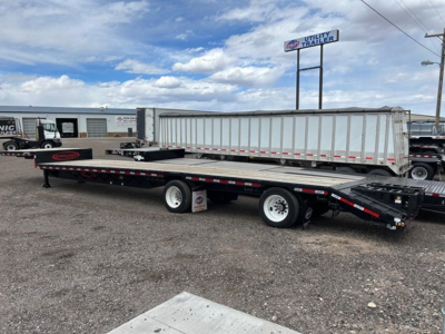 Trailer in a lot showing a long steel drop deck trailer with dual rear axles and a gooseneck area.