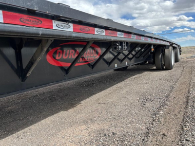Trailer side showing DURA-HAUL logos and black drop deck with red oval branding, wheels visible at rear, flat open landscape.