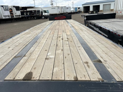 Flatbed trailer deck showing wooden planks with metal edge channels, used condition, long stretch of board decking, surrounding trailers and yard in background.