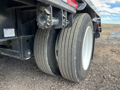 Dual rear tires of a drop deck trailer with black frame and white wheel rims, gravel ground, clear sky.
