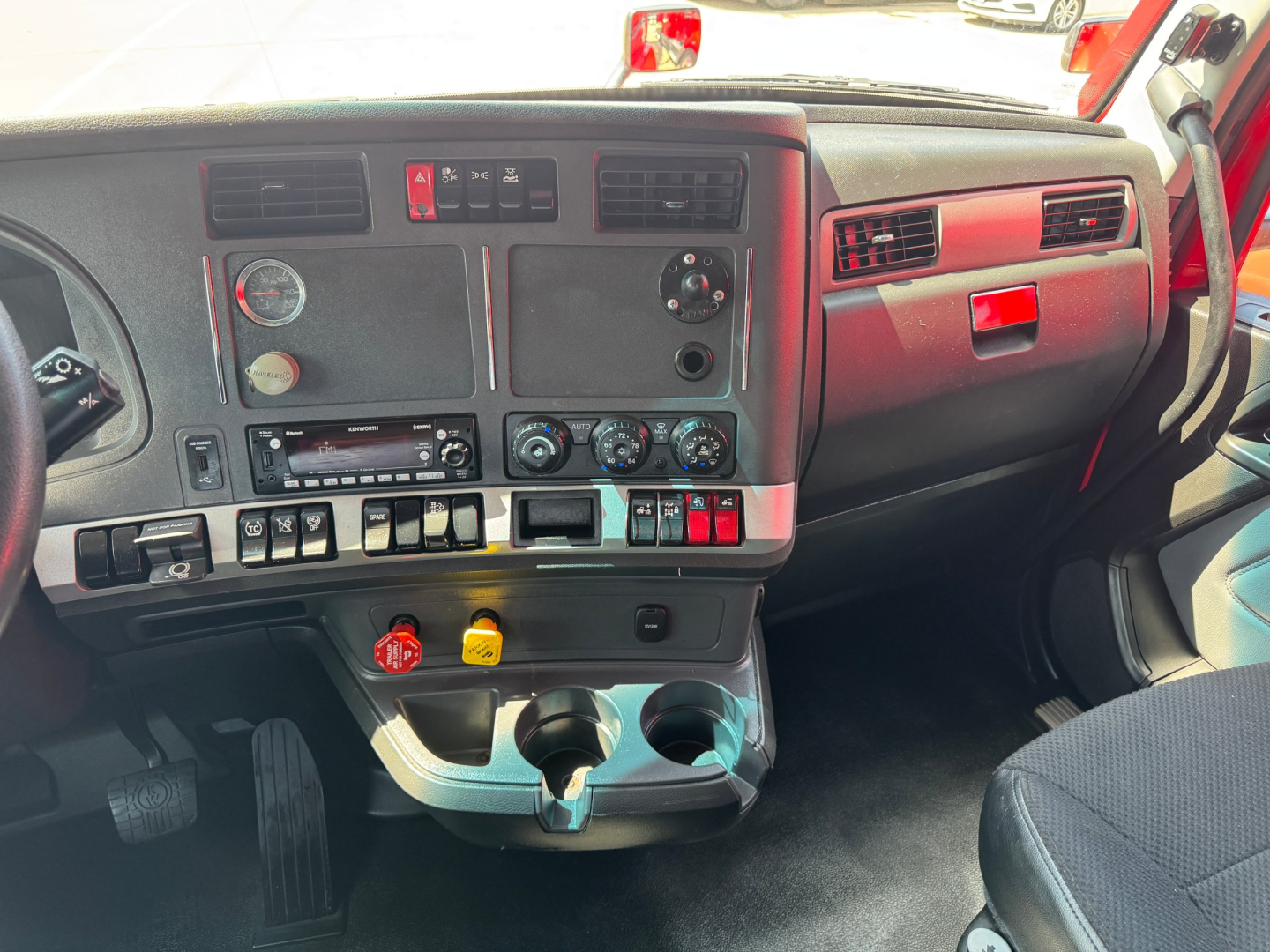 Kenworth T680 cab interior dashboard view showing steering column, control switches, HVAC vents, gauges, cup holders and glove compartment.