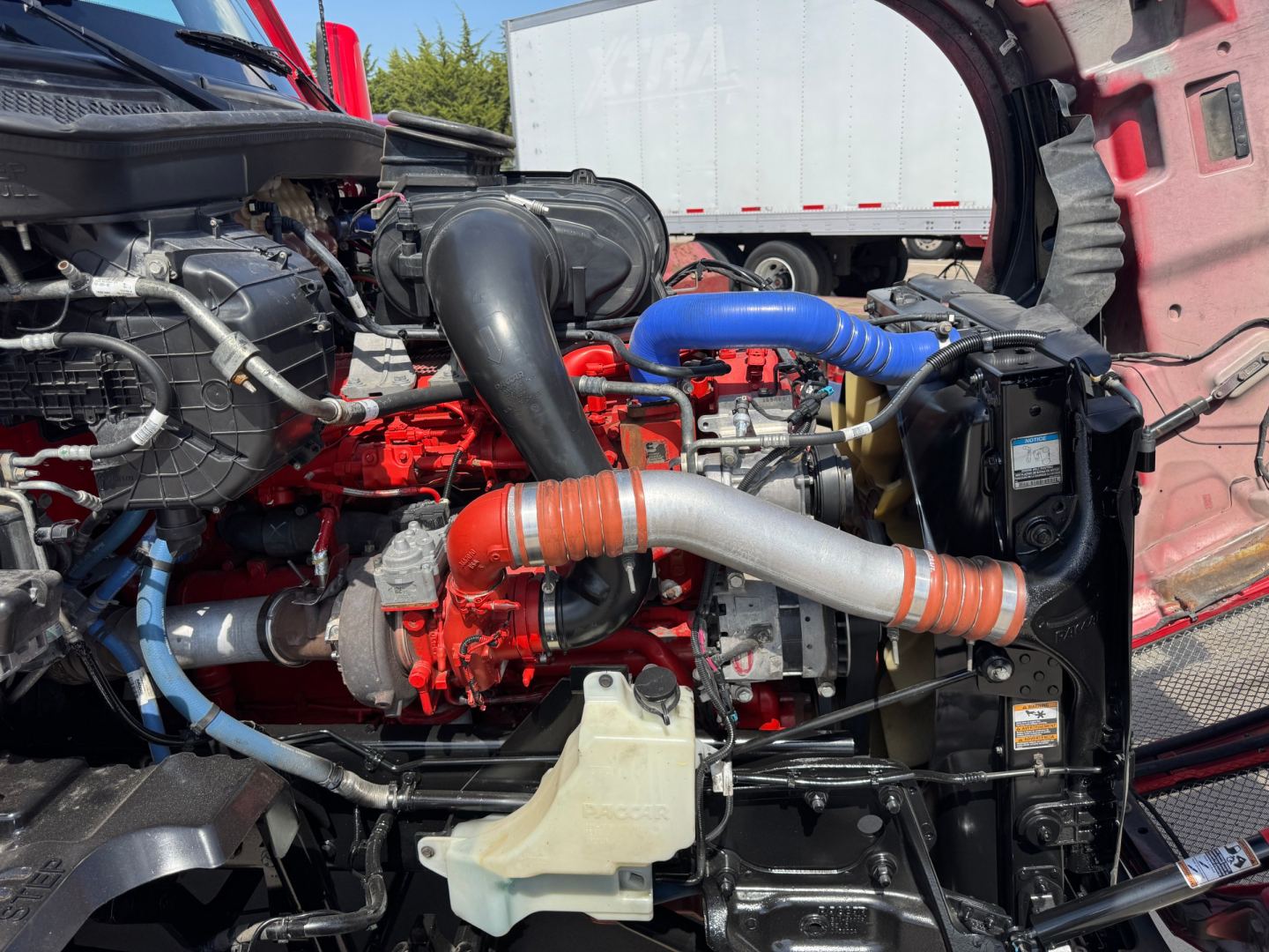 Kenworth T680 engine bay showing red engine block, blue and orange intercooler hoses, black radiator, coolant reservoir, and various metal tubing.