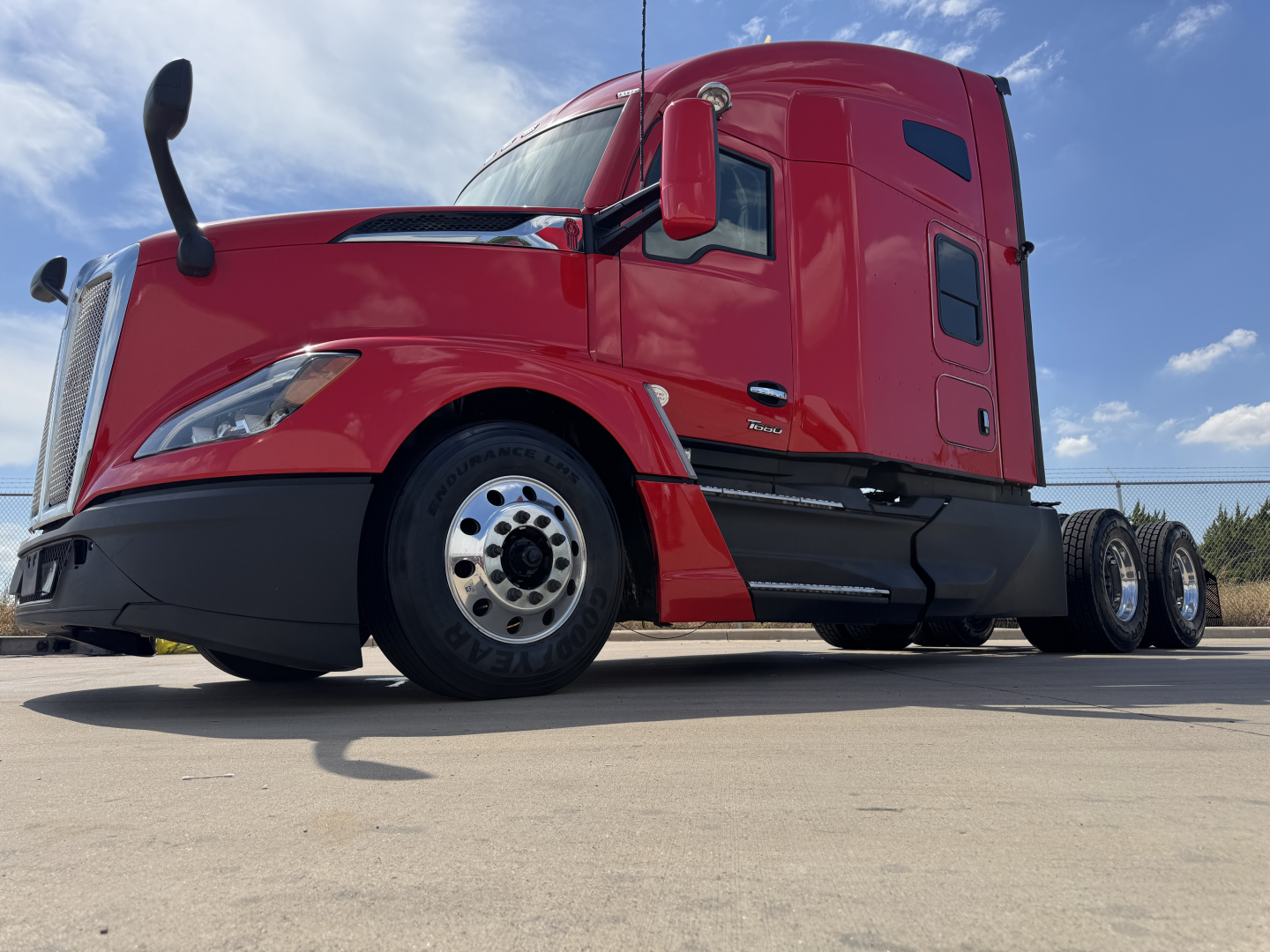 Kenworth T680 tractor in red exterior, low-angle view, showing front left wheel, aerodynamic fenders, side exhaust, and sleeper cab.