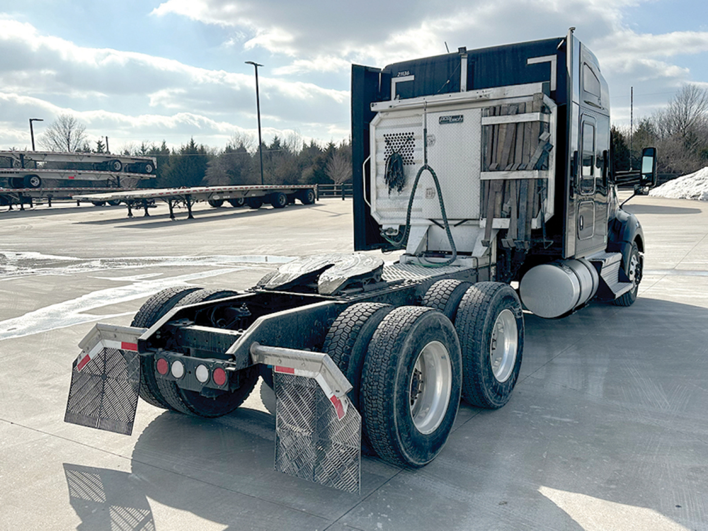 Kenworth T680 tractor in a parking lot rear-three-quarter view showing drivetrain, five rear wheels, and cooling unit with hoses.