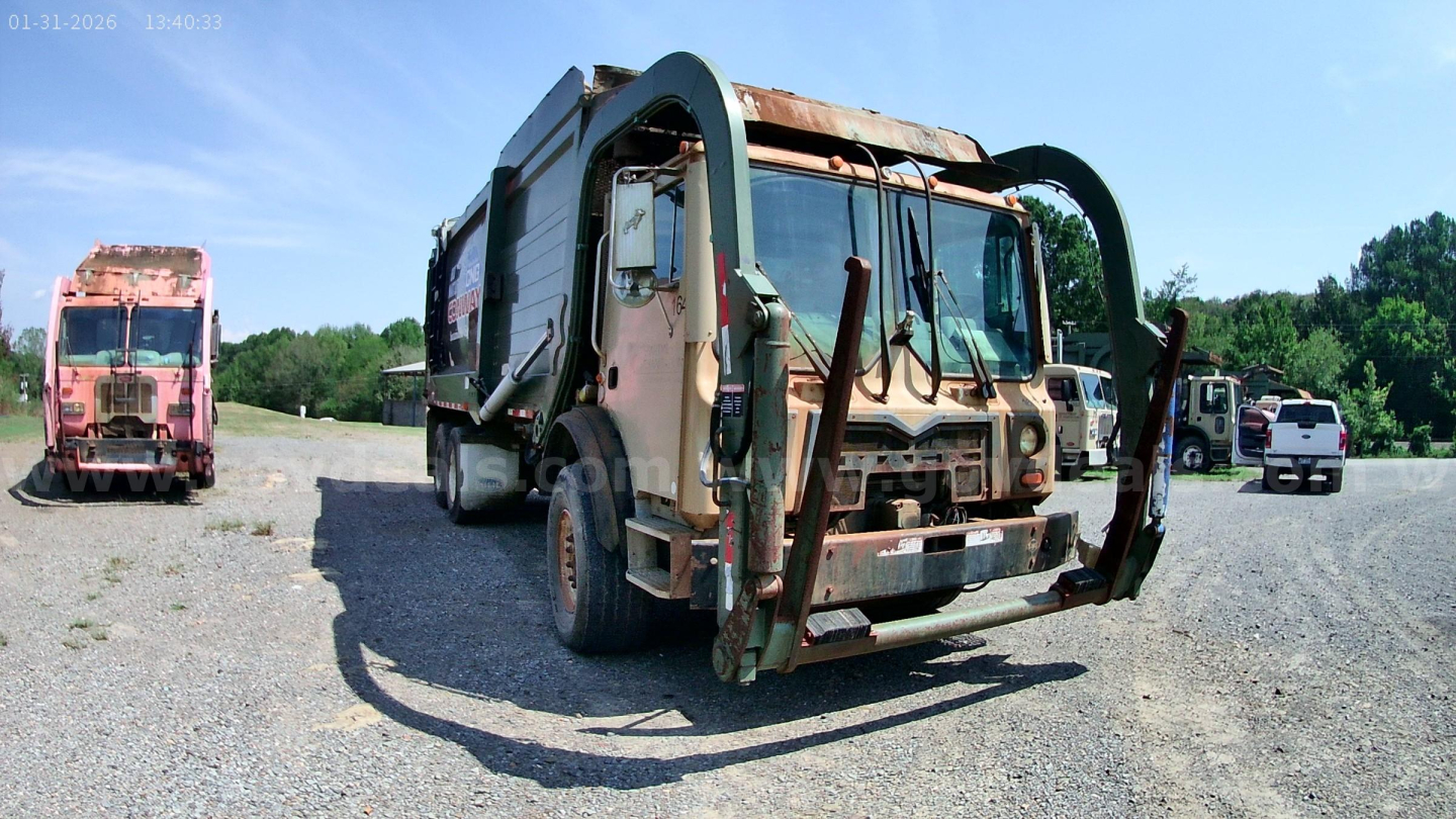 Front view of a beige Mack MRU613 refuse truck with green lifting arms, rusty bumper, and another pink truck in the background; clear sky.