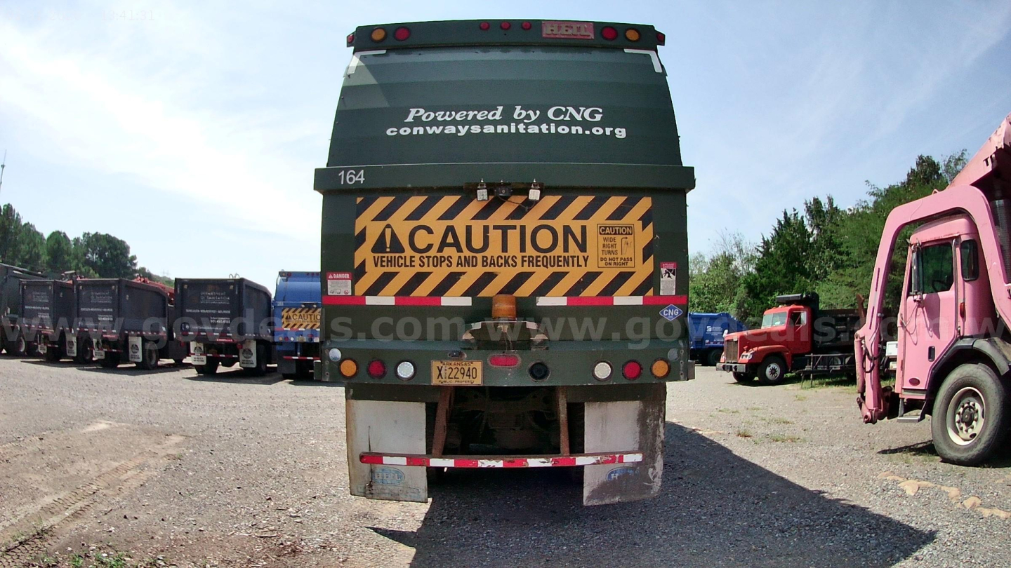 Rear view of a 2015 Mack MRU613 truck showing caution stripes and a waste vehicle graphic, with other trucks in a lot.