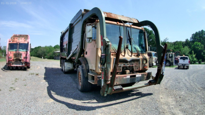 Front view of a beige Mack MRU613 refuse truck with green lifting arms, rusty bumper, and another pink truck in the background; clear sky.