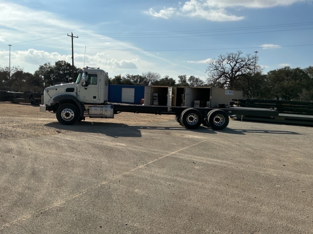 Truck cab and long flatbed crane chassis in an empty lot showing side view with Mack GR64B cab and crane equipment.