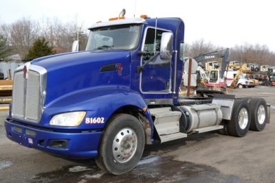 Kenworth T660 conventional daycab tractor in blue, side view showing front grille, headlights, fuel tanks, and dual rear axles with a hook lift and equipment in background.
