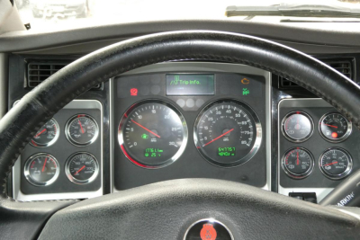Dashboard of a Kenworth truck showing multiple gauges and warning lights, including speedometer, tachometer, and digital readout, with steering wheel in foreground.