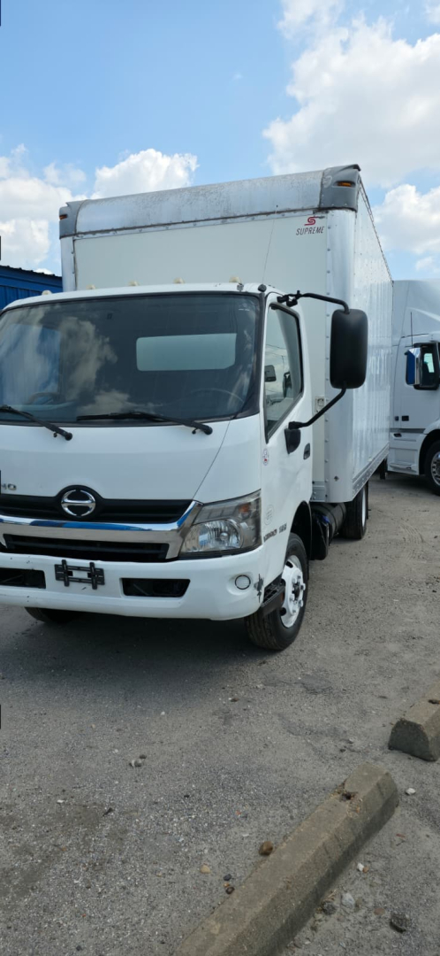 White 2019 Hino box truck front-left view, cab with Hino emblem, large box cargo area, side mirrors, daytime sky.