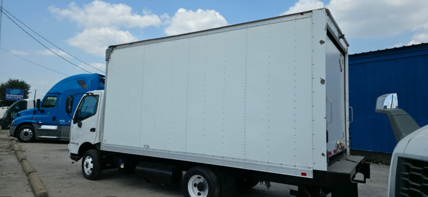 Box truck side view showing a white cargo box on a Hino 155 with a blue cab and another blue truck in background